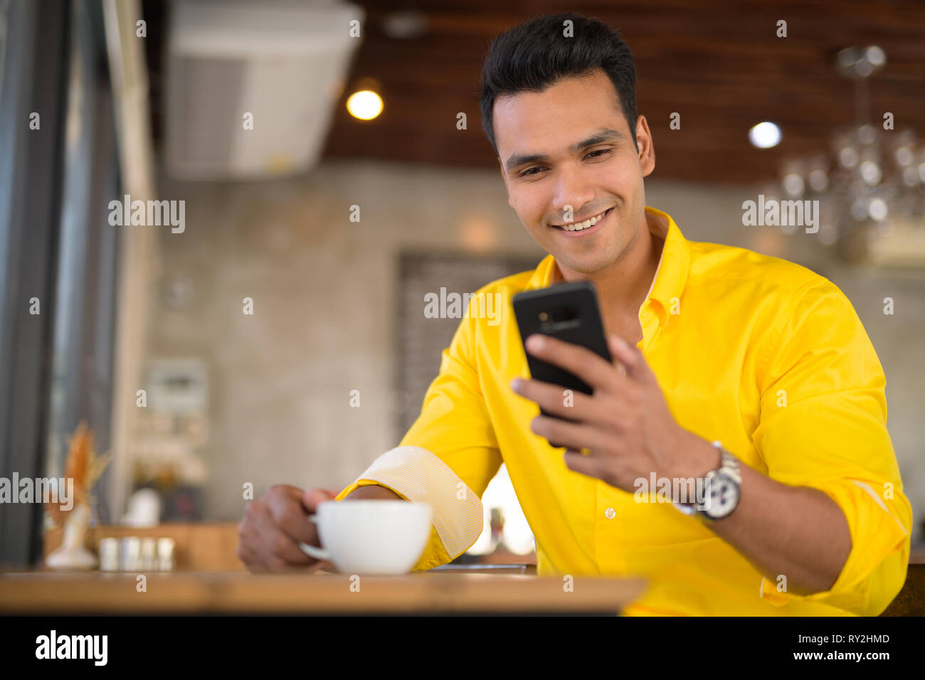 Giovani felici uomo indiano tramite telefono e seduta dalla finestra al cafe Foto Stock