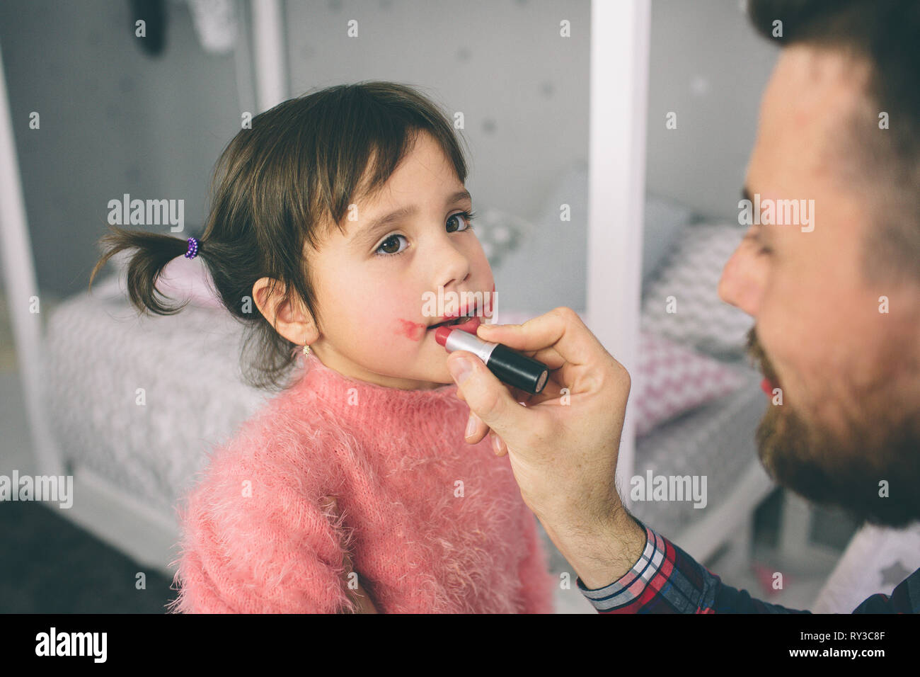 Incantevole piccola figlia ed il suo bel giovane papà sta giocando insieme in stanza per bambini. Ragazza sta facendo il suo papà un trucco. Foto Stock