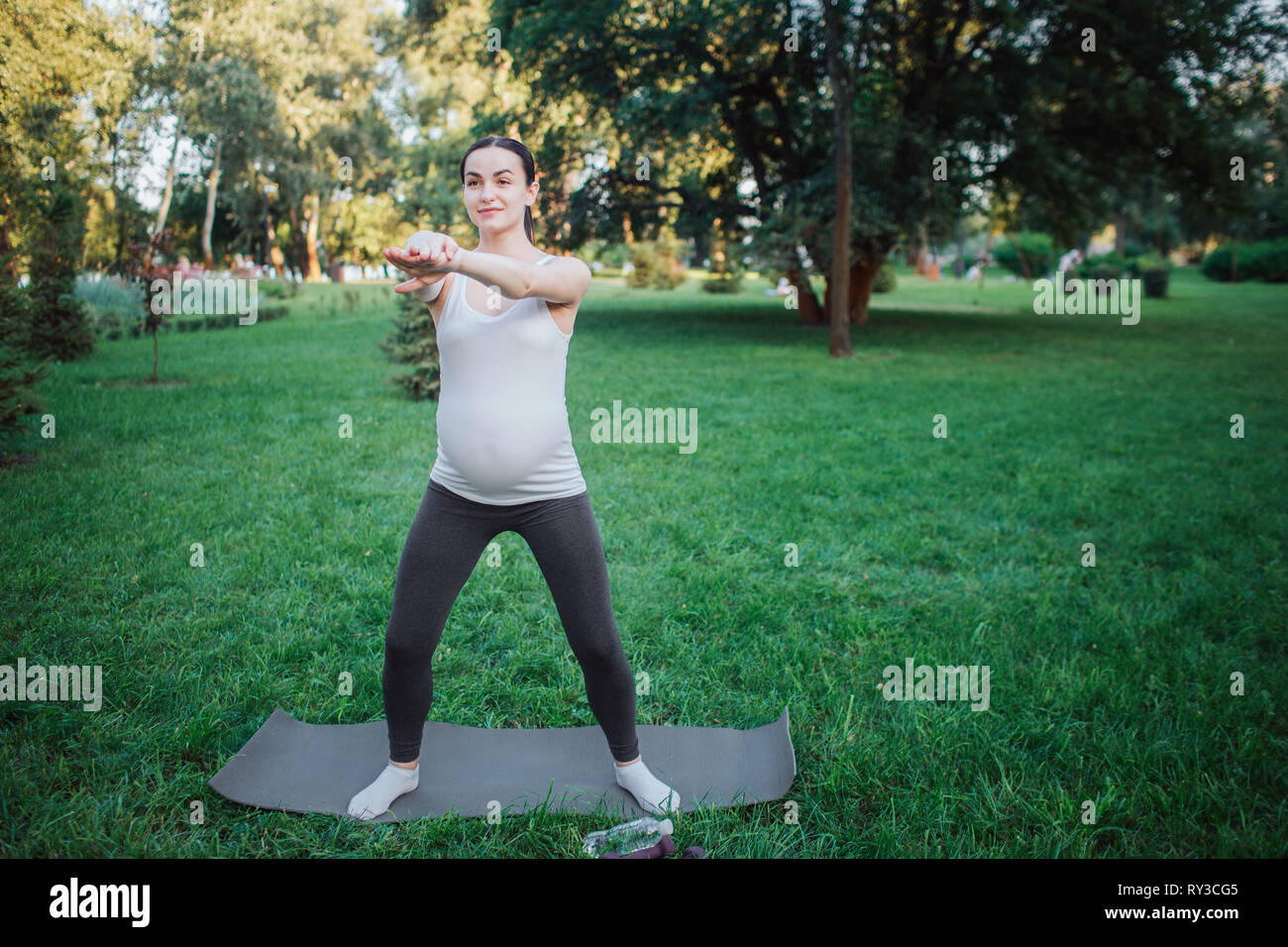 Giovane donna incinta stand sul compagno di yoga al di fuori nel parco. Ella ha tratto le braccia e guardare avanti. Guarda il modello concentrato. Foto Stock