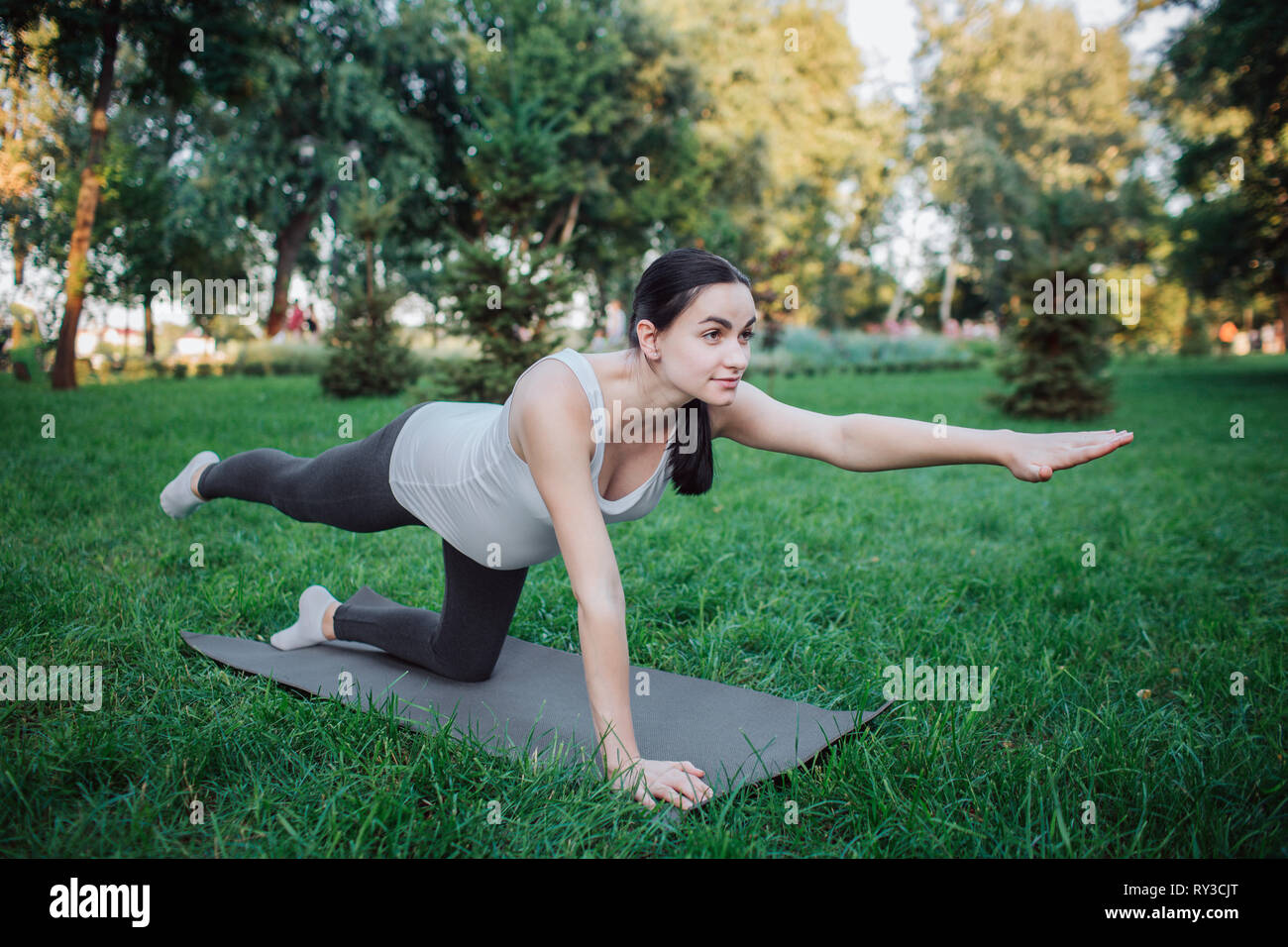 Giovane donna incinta stand sulle ginocchia e di esercitare sul compagno di yoga nel parco. Ella lo stiramento delle gambe e braccia. Concentrato e grave donna di guardare avanti. Foto Stock