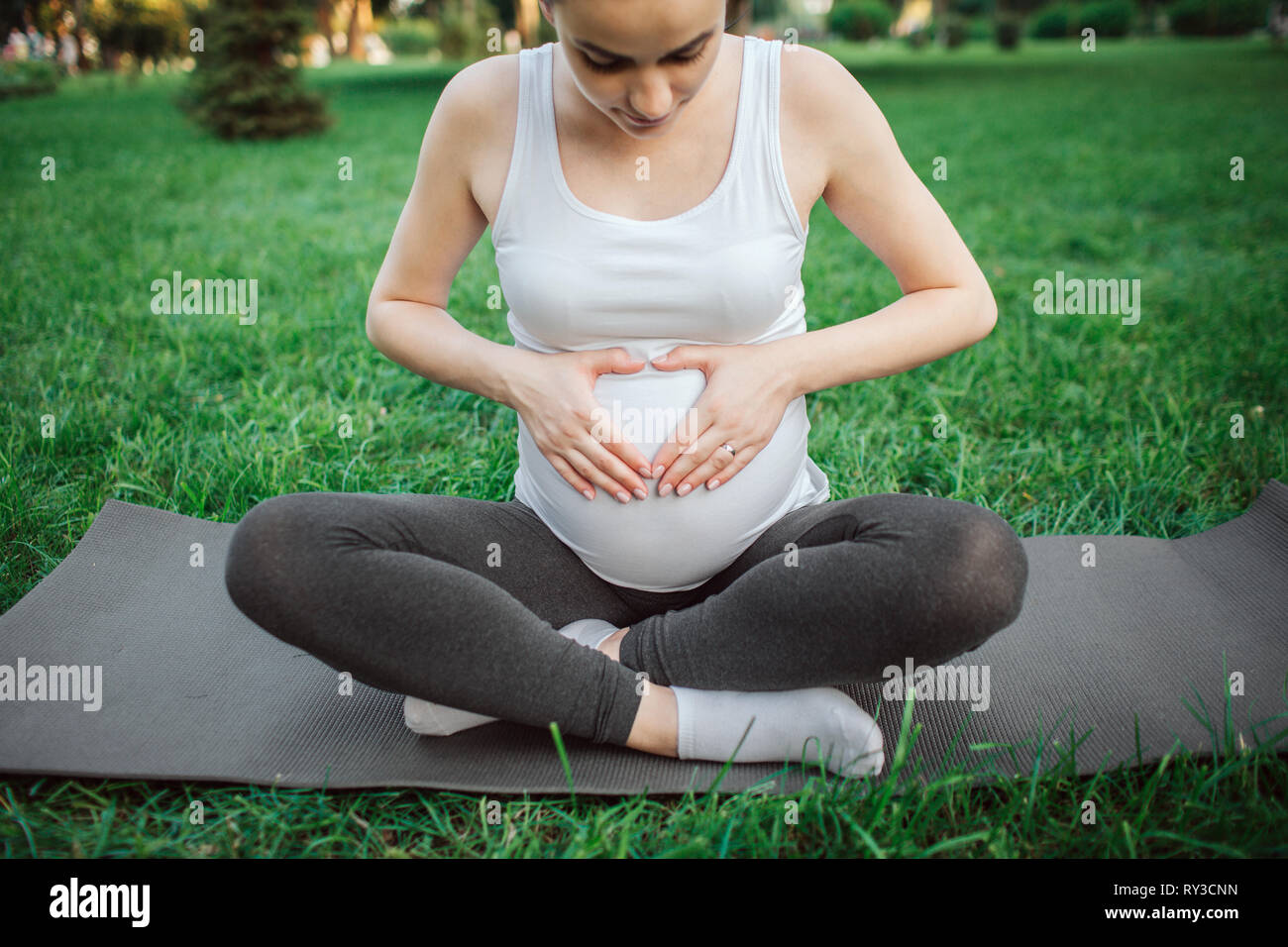 Giovane donna incinta sit in lotus pongono sul compagno di yoga nel parco. Lei guarda la pancia e tenere le mani in forma di cuore. Foto Stock