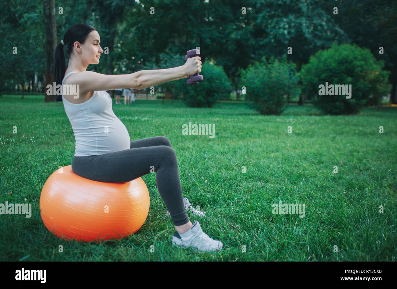 Immagine della bella giovane donna incinta sedersi sul fitness arancione palla in green park. Ella esercizio con manubri. Model look concentrato. Foto Stock