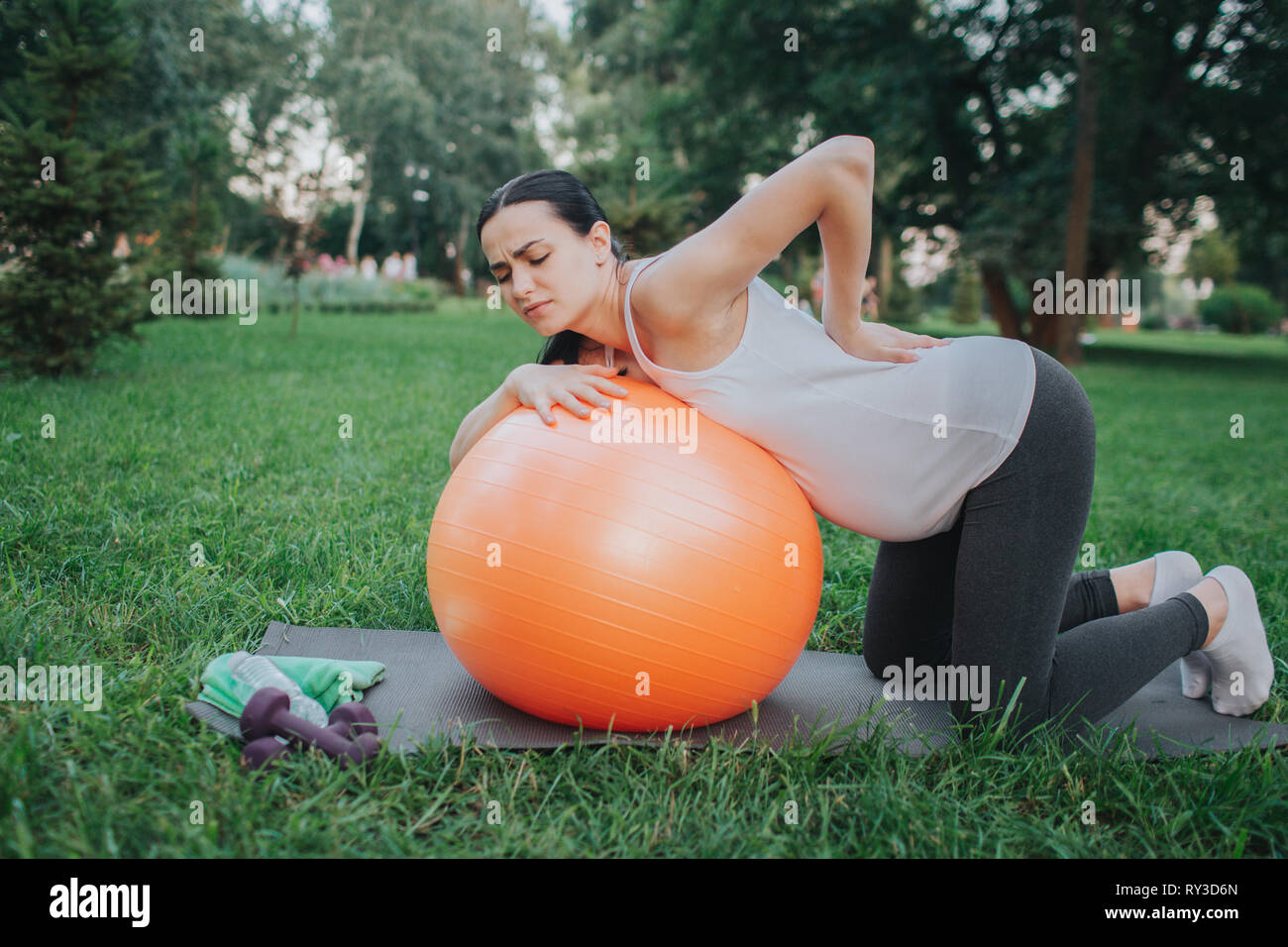 Giovane donna incinta suffeer dal dolore nel retro. Stand ella in ginocchia nel parco. Modello di magra arancione palla fitness. Foto Stock