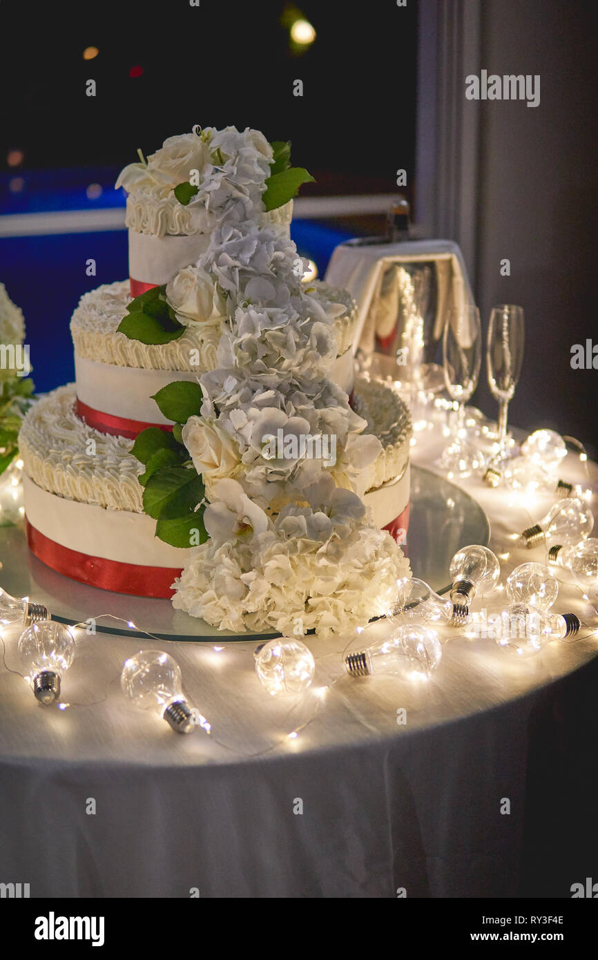 A tre livelli di bianco torta di nozze con decori di rose su un tavolo decorato con candele bianche e lampadine. Formato verticale. Foto Stock