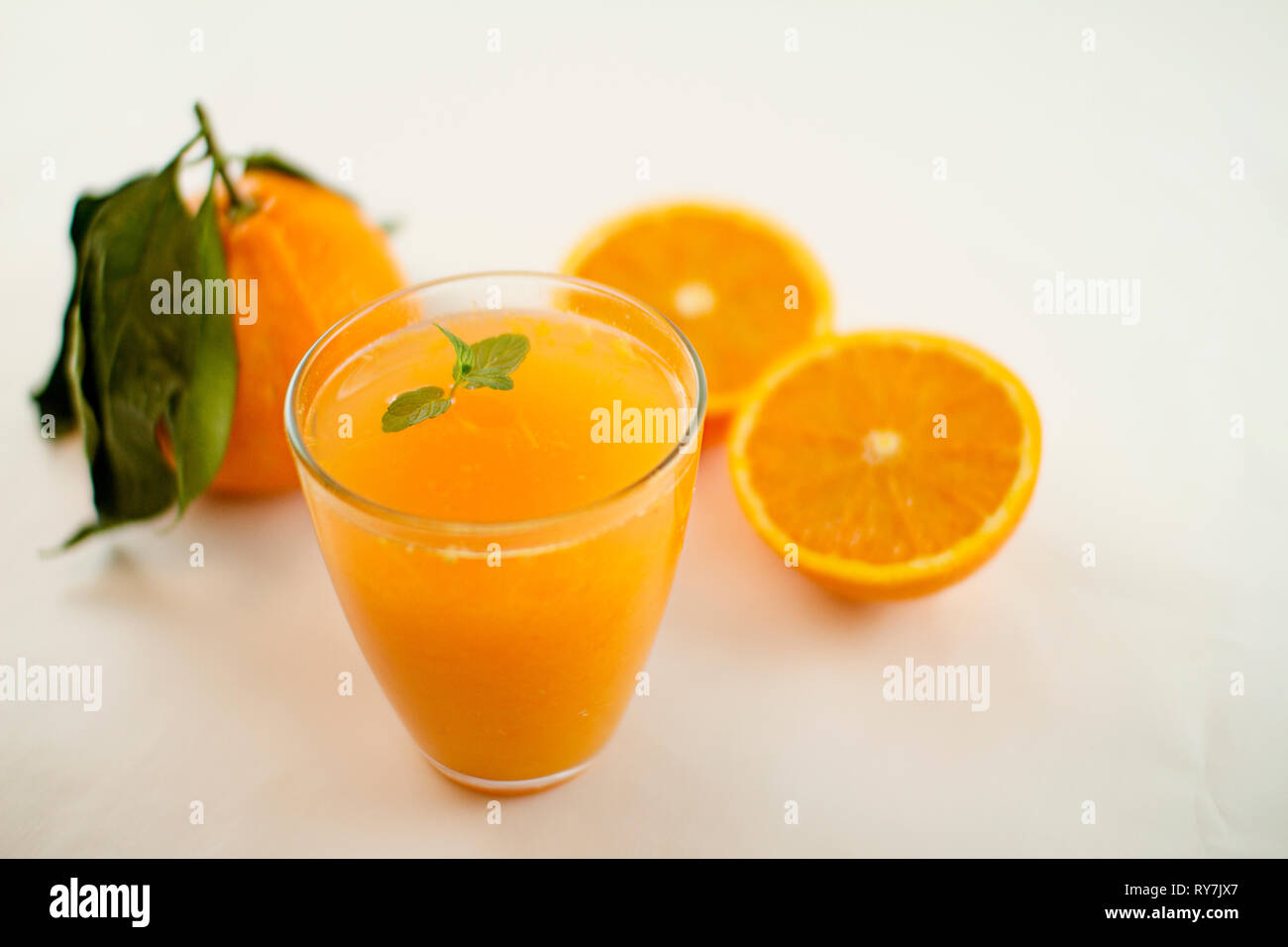 Un invitante bicchiere pieno di succo di arancia con una fresca foglia di menta che galleggia. Sullo sfondo un arancione diviso in due e un intero uno con foglie su un Foto Stock