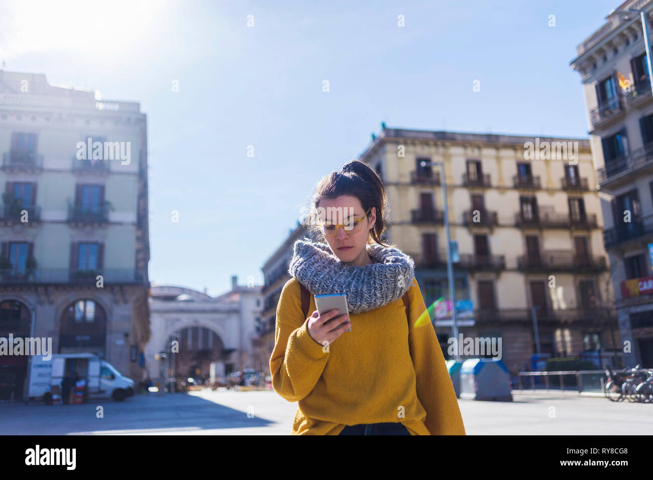 Moda turista femminile di indossare un abbigliamento caldo utilizzando smart phone mentre in piedi in città durante la giornata di sole Foto Stock