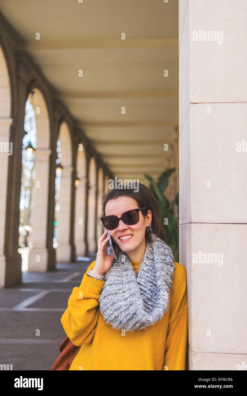 Ritratto di sorridente turista femminile di indossare un abbigliamento caldo e occhiali da sole rispondendo alle smart phone mentre in piedi in corridoio Foto Stock