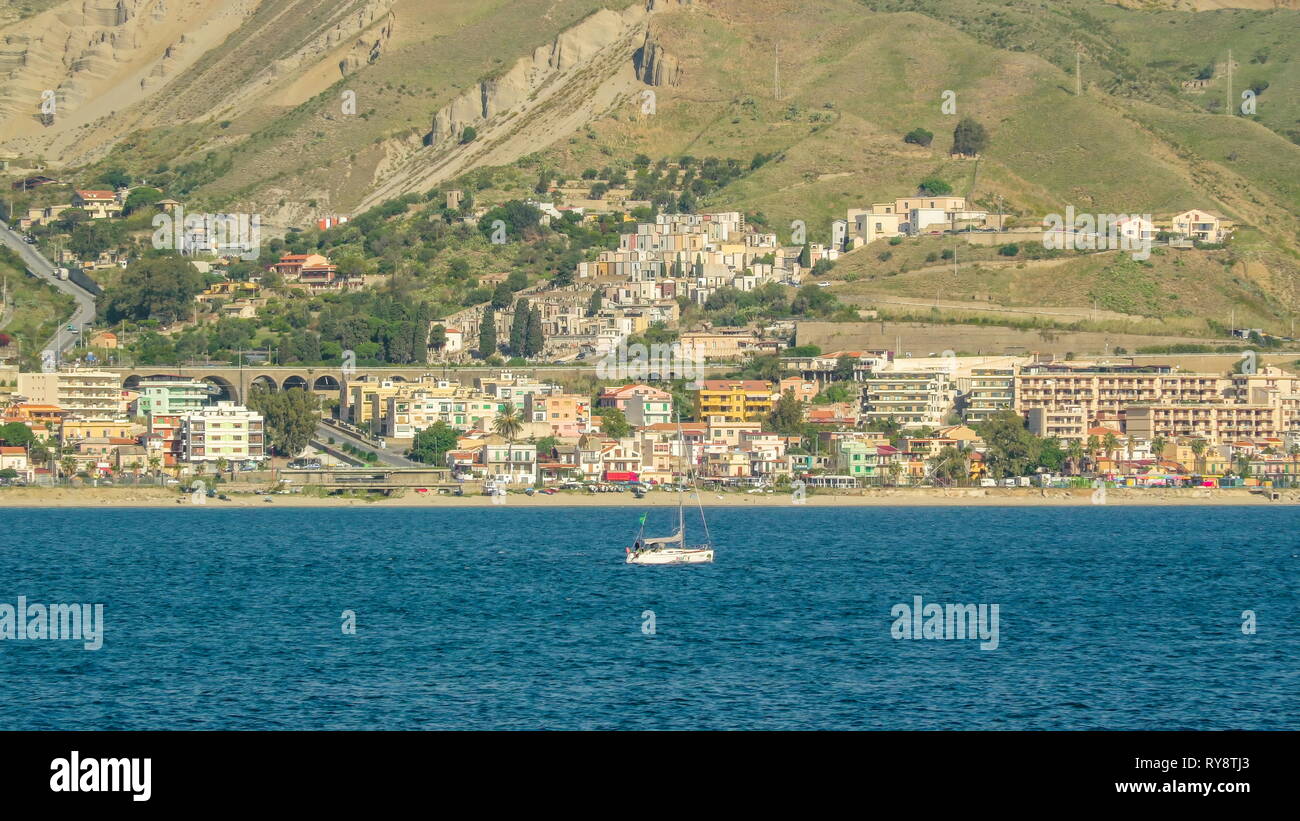 Una piccola barca a vela bianca in mezzo al mare e la vista delle case residenziali sulla montagna a Villa San Giovanni in Italia Foto Stock