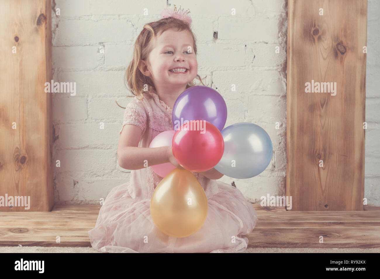 Bambina in rosa pastello abito e fascia di corona tenendo in color pastello e vibrante baloons, legno solido background mantelpiece, tonica, il fuoco selettivo Foto Stock