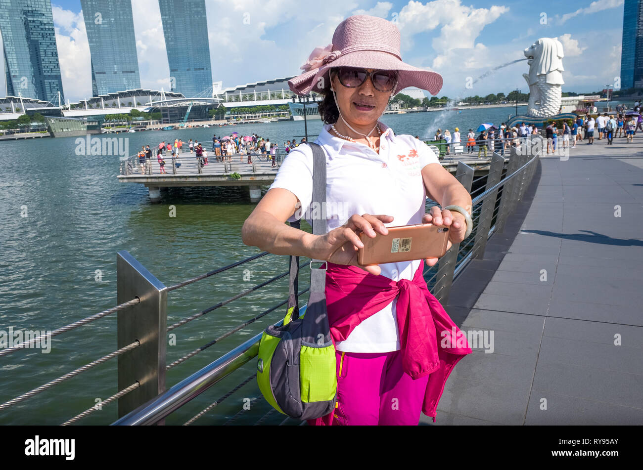 Coreano donna turistica in rosa cappello, tenendo selfie con statua Merlion e Marina Bay Sands, Singapore Foto Stock