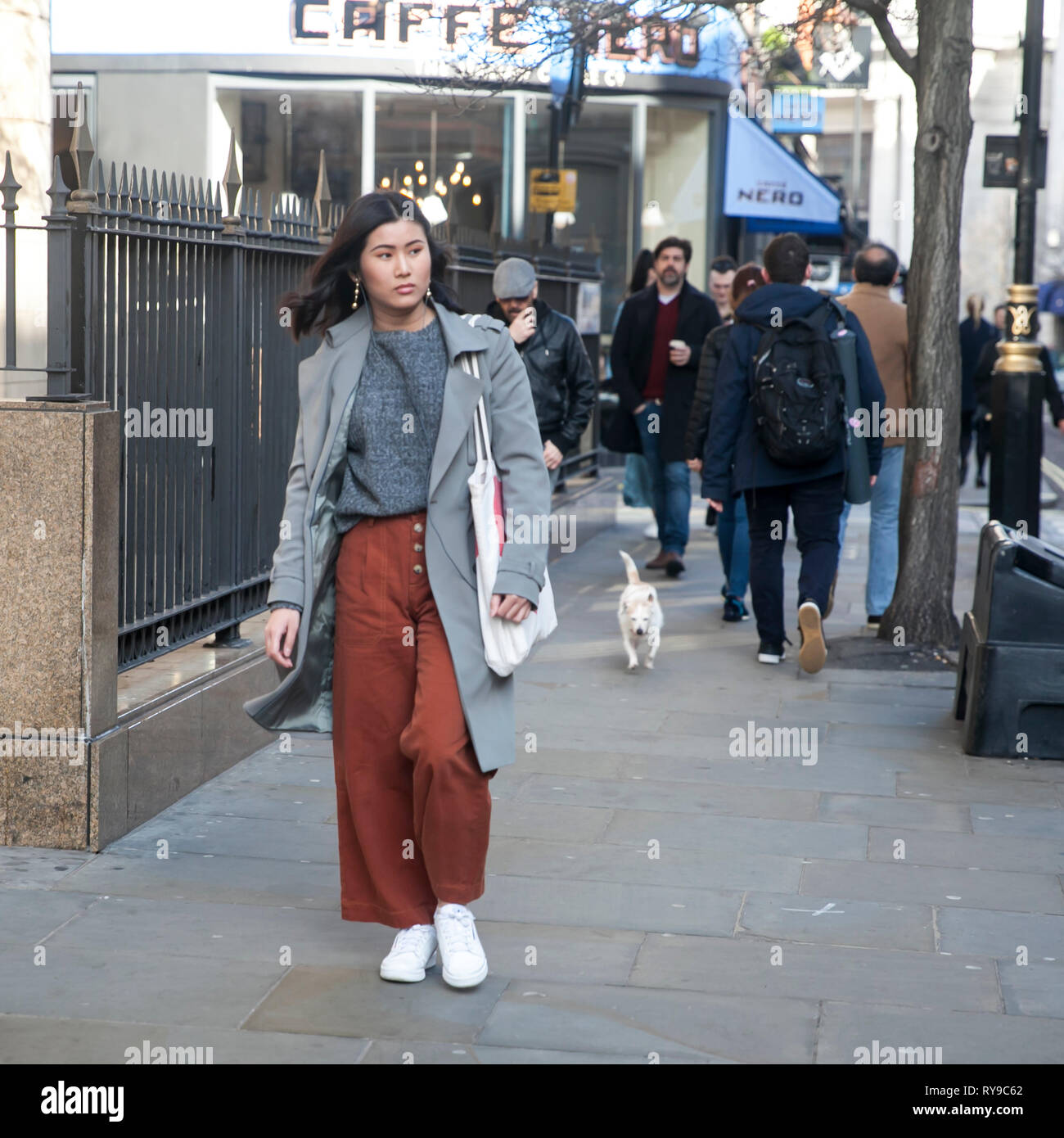 Londra - 17 febbraio 2019: Ragazza in un cardigan grigio e marrone e pantaloni svasati cammina giù per la strada. Ragazza in un cardigan grigio e marrone e pantaloni svasati Foto Stock