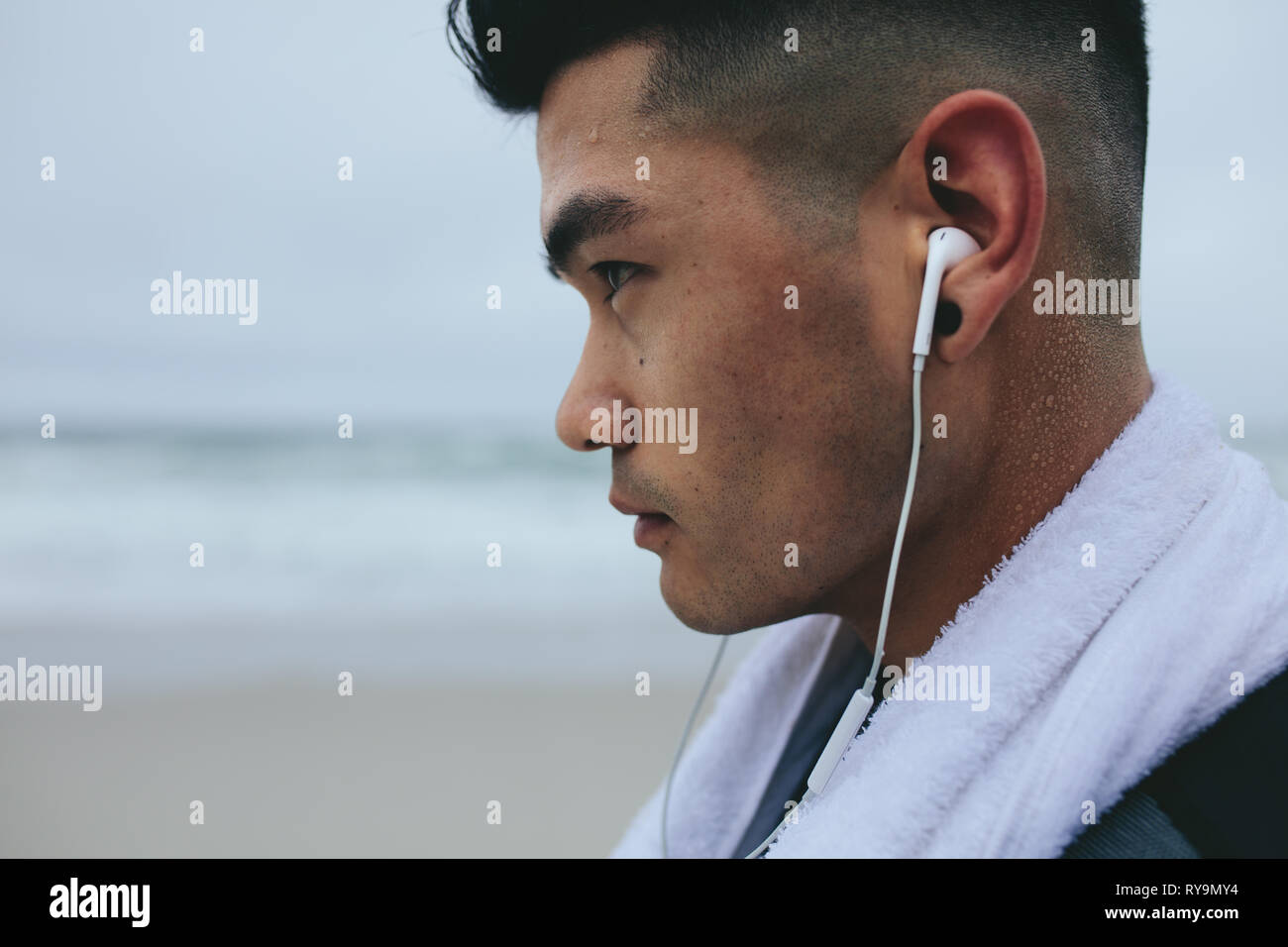 Chiusura del giovane uomo asiatico con auricolare e l'asciugamano intorno al suo collo. Maschio modello fitness ascoltare musica con gli auricolari tenendo il resto dall'esercizio. Foto Stock