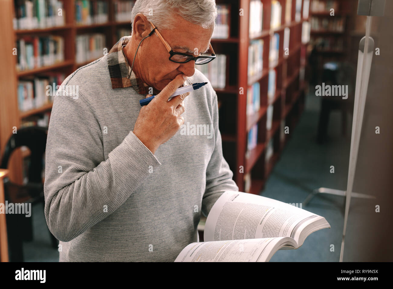 Professore universitario di insegnamento in classe in piedi con un libro e un marcatore in mano. Senior uomo la lettura di un libro con la concentrazione in piedi in aula. Foto Stock