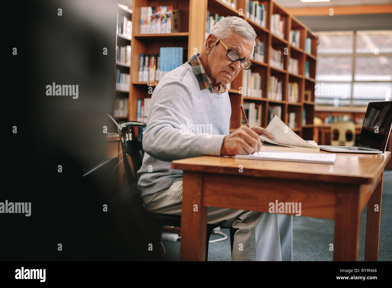 Senior uomo studiare in aula. Uomo anziano iscritto in un libro seduti in classe con un computer portatile nella parte anteriore. Foto Stock