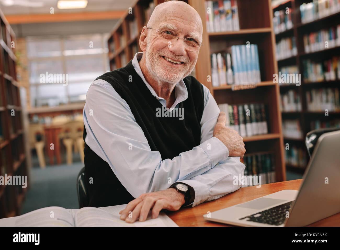 Senior sorridente uomo seduto in una biblioteca con i suoi libri e computer portatile sul tavolo. Allegro senior uomo seduto in biblioteca e apprendimento. Foto Stock
