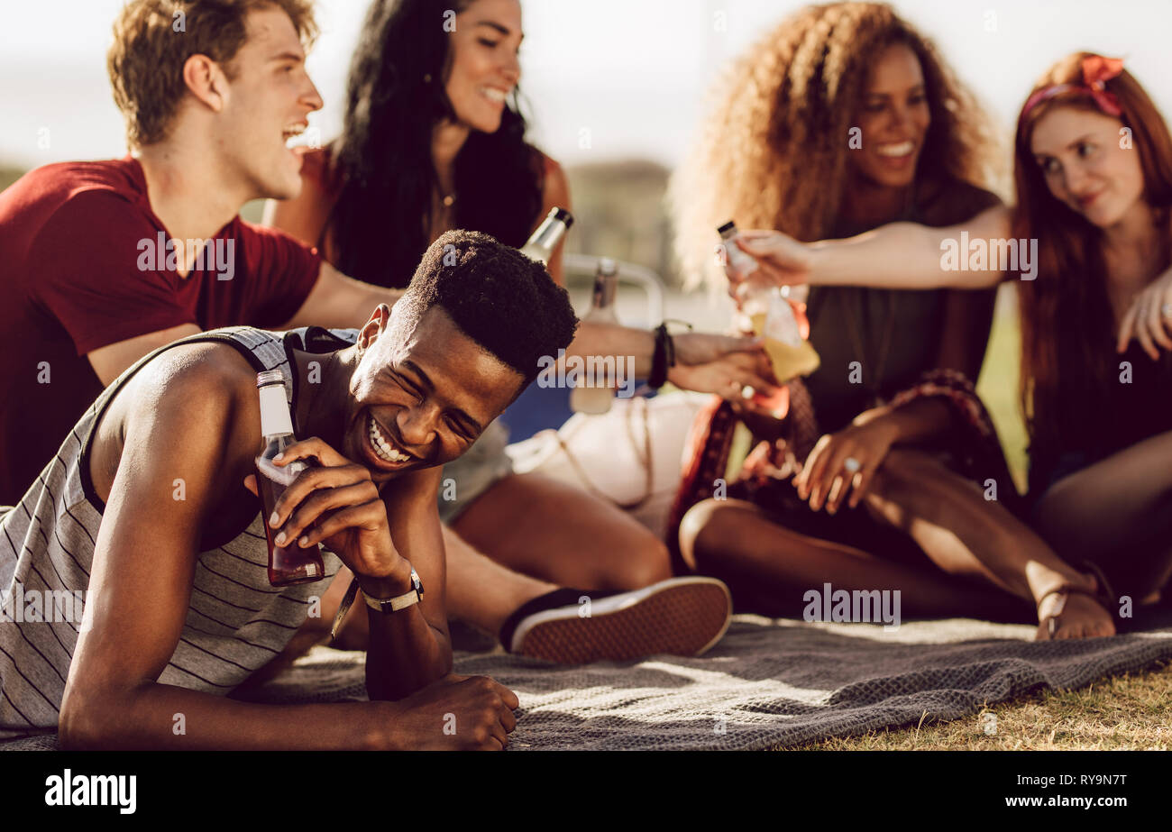Allegro giovane africana ragazzo seduto con gli amici all'aperto e ridere. L'uomo godendo summer weekend con gli amici. Foto Stock Allegro giovane africana ragazzo seduto con gli amici all'aperto e ridere. L'uomo godendo summer weekend con gli amici. Foto Stock