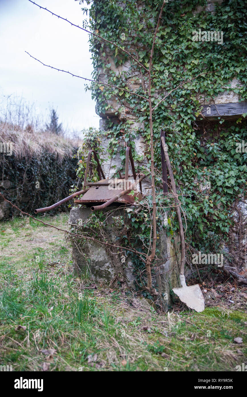 Vecchie e arrugginite carriola e la pala su una parete di una casa ricoperta con poison ivy nel villaggio abbandonato Slapnik nella regione Goriška Brda, Slovenia. Foto Stock