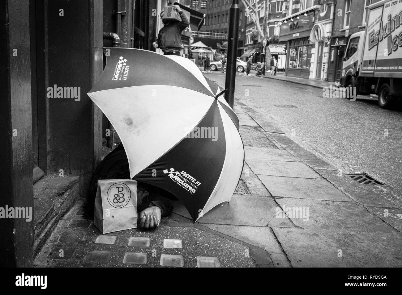 Fotografia di strada in bianco e nero a Londra: Uomo senza tetto che usa un grande ombrello per il riparo sulla via centrale di Londra. REGNO UNITO Foto Stock