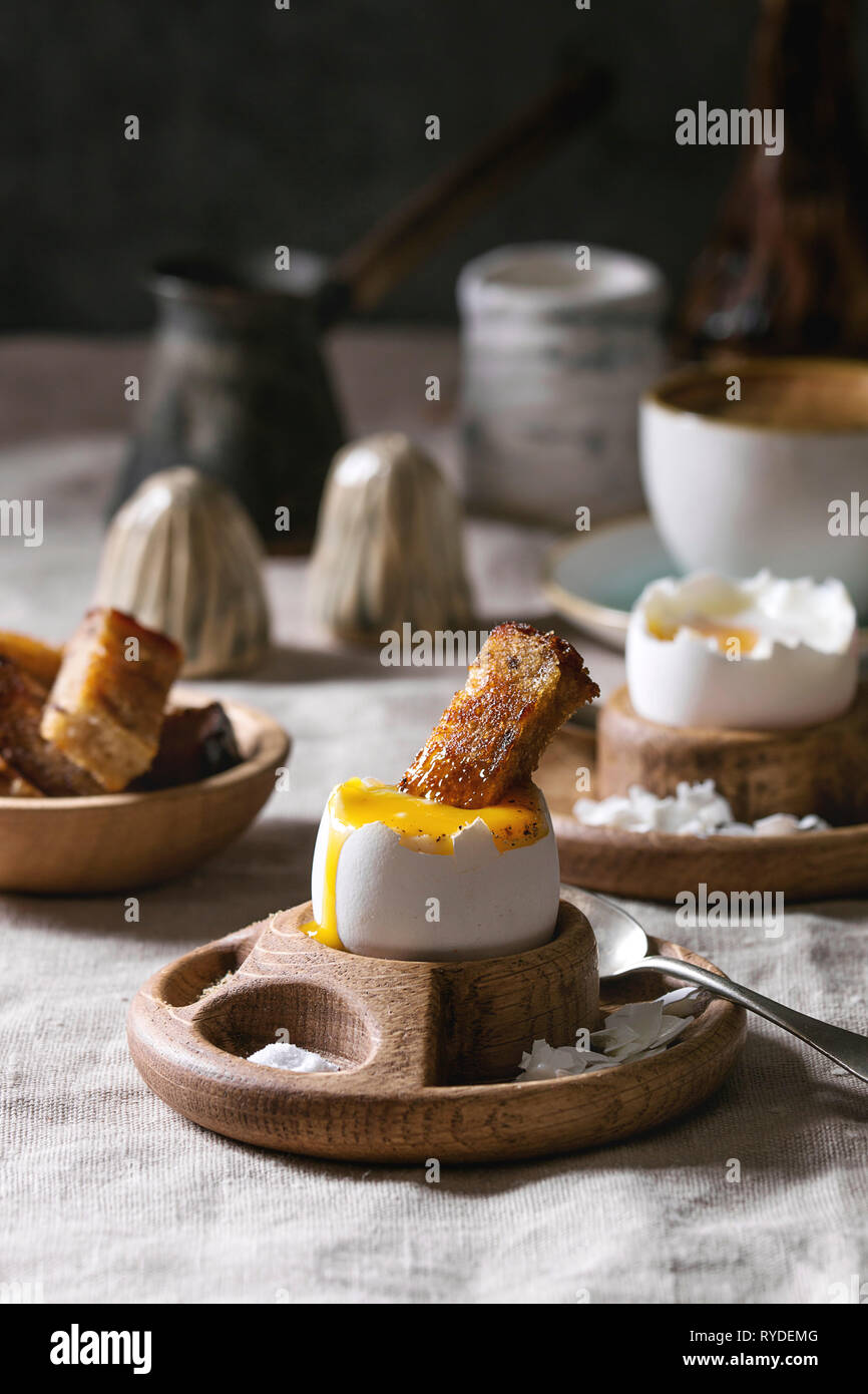 La prima colazione con la tazza di caffè e uova sode, servita in legno tazza di uova con sale e pepe e il pane tostato, brocca di crema su tovaglie di lino. Foto Stock