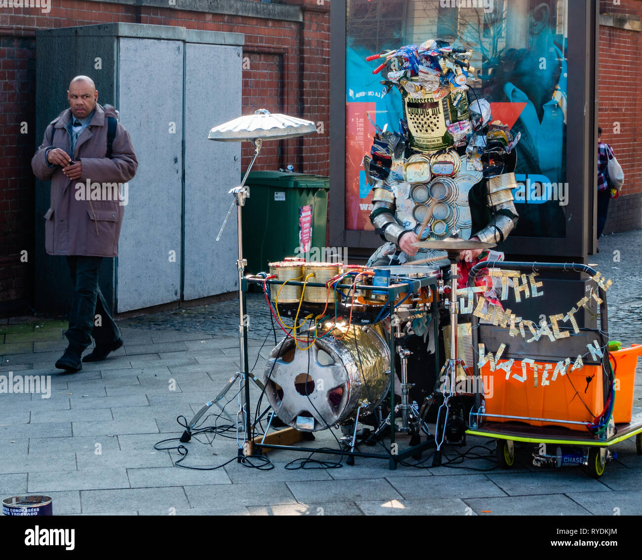Uomo che cammina davanti a un suonatore ambulante Il Junkoactive Wasteman e il suo Tinphonia one man heavy metal band di oggetti riciclati in Bristol City Centre Regno Unito Foto Stock