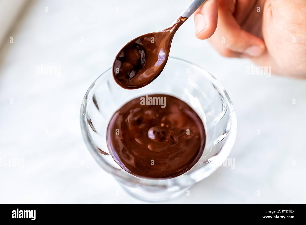 Primo piano della mano che tiene il cucchiaio mescolando sotto agitazione uniforme di liquido fuso a caldo di crema di cioccolato sciroppo di glassa salsa in piccola ciotola di vetro piatto Foto Stock
