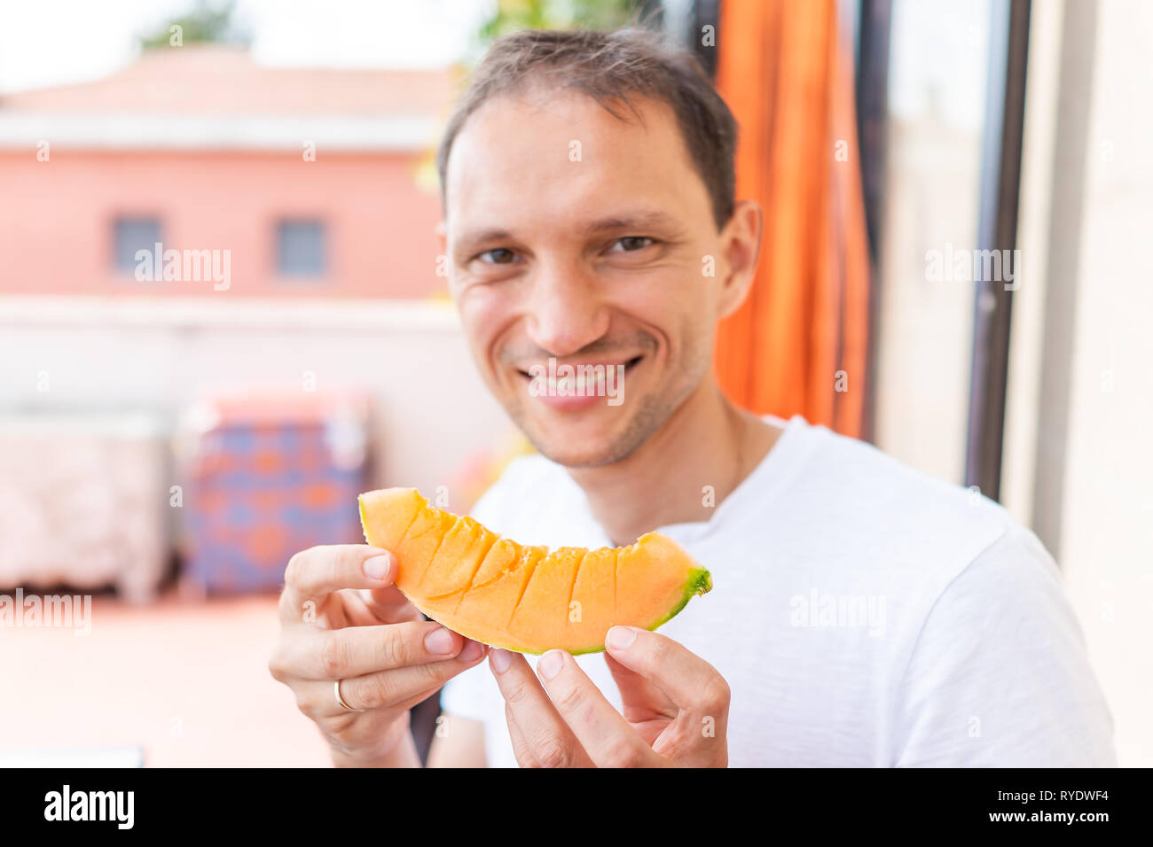 Giovane uomo felice in bianco estate shirt sorridente mangiare vibrante colorato arancione melone fette di melone al di fuori in Italia villa soleggiata la luce solare Foto Stock