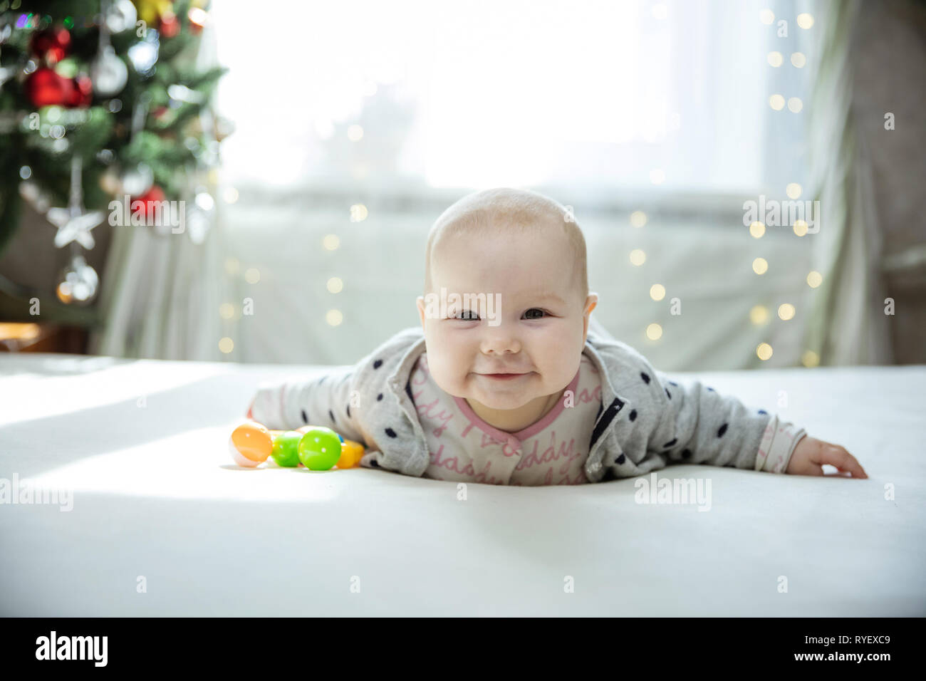 Carino Baby girl sdraiato sul letto di casa e sorridente. Albero di natale in background. Foto Stock