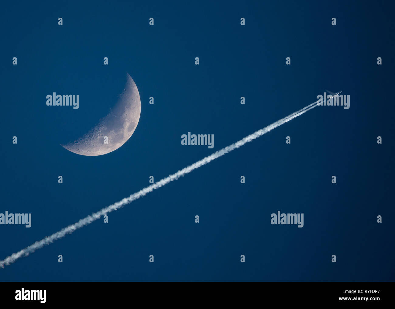 Un ad alta quota con jet contrails passa da una falce di luna in un cielo blu scuro al tramonto. Foto Stock