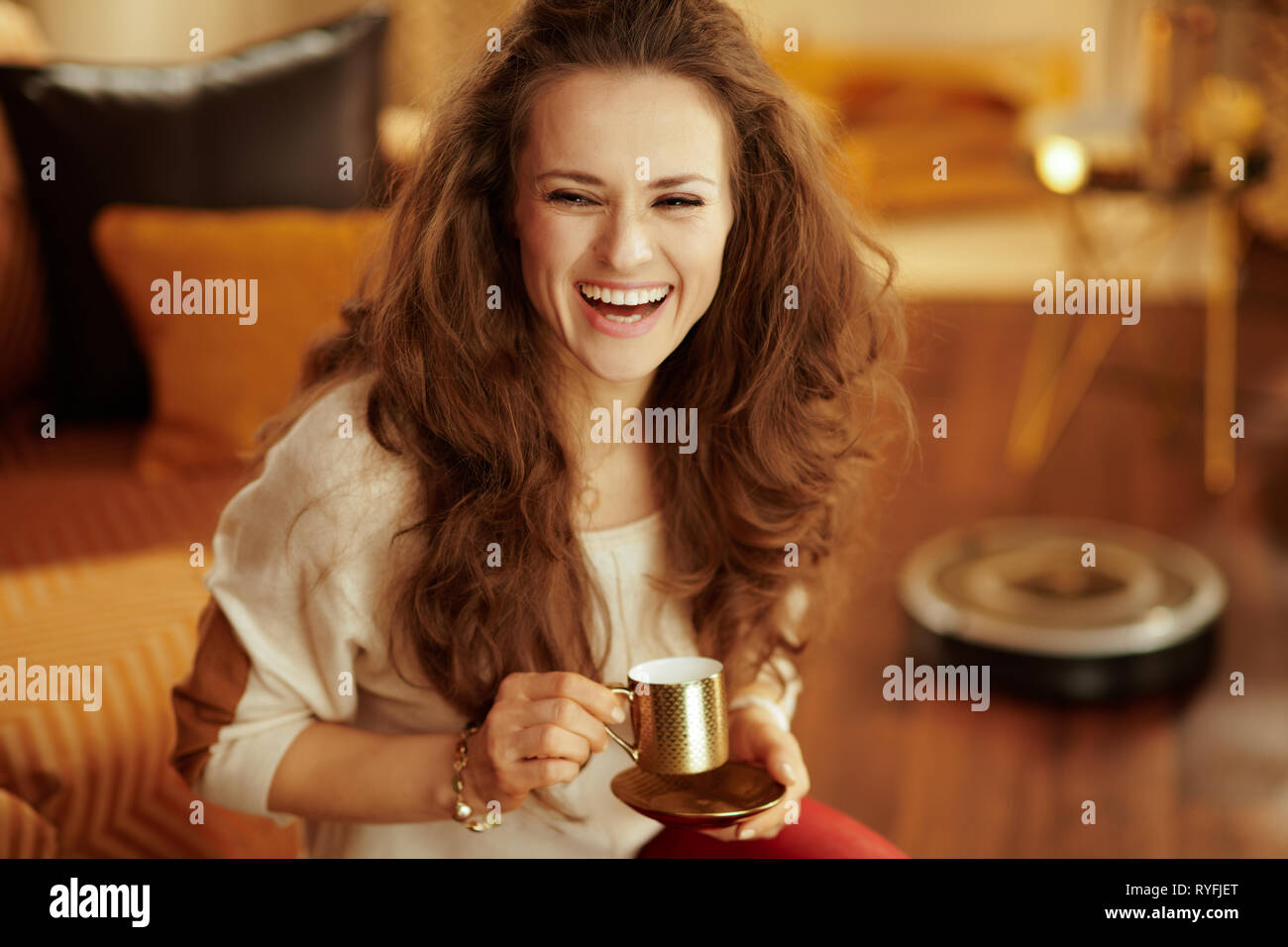 Sorridente giovane donna con lunghi capelli bruna nel soggiorno moderno di bere il caffè mentre il robot a pulire il pavimento. Foto Stock