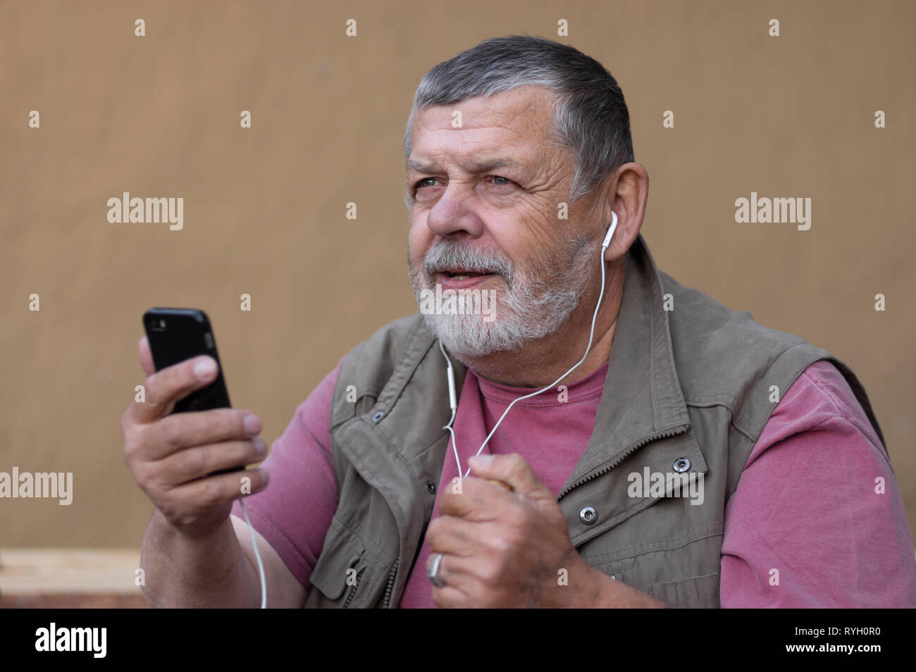 Senior barbuto uomo che ascolta musica dal cellulare tramite le cuffie mentre è seduto all'esterno della parete di argilla Foto Stock