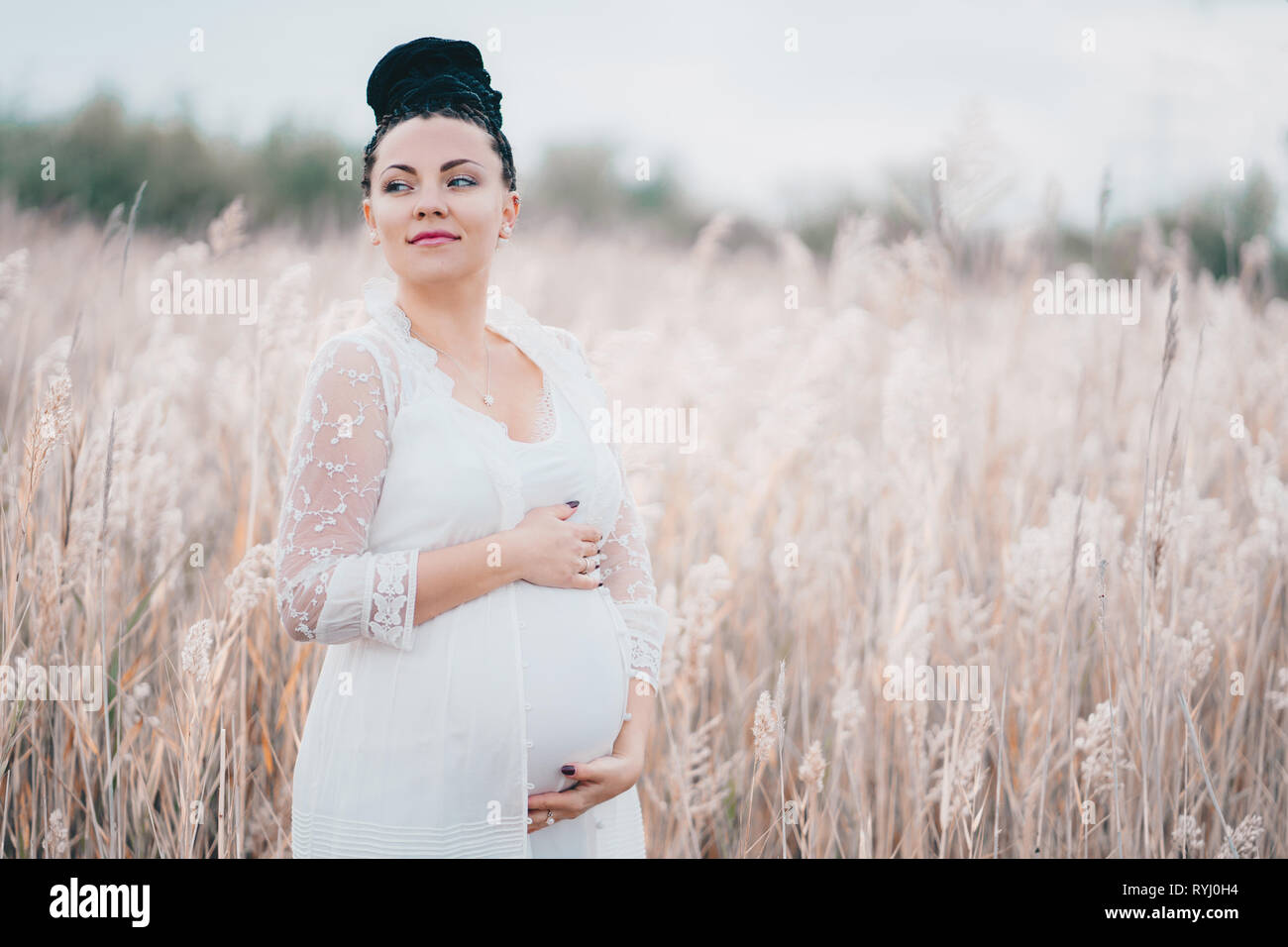 Incinta bella donna in pizzo bianco boho abito in piedi in canne e tenendo il suo ventre Foto Stock