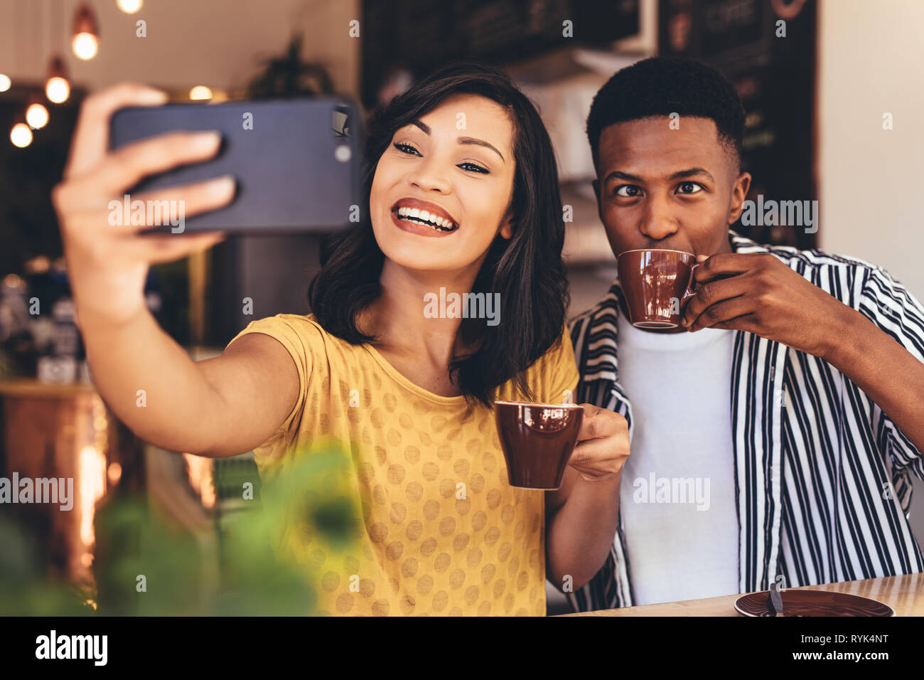 Donna prendendo selfie con amico rendere divertente espressioni mentre si beve il caffè. Amici prendendo selfie con smart phone al cafe. Foto Stock