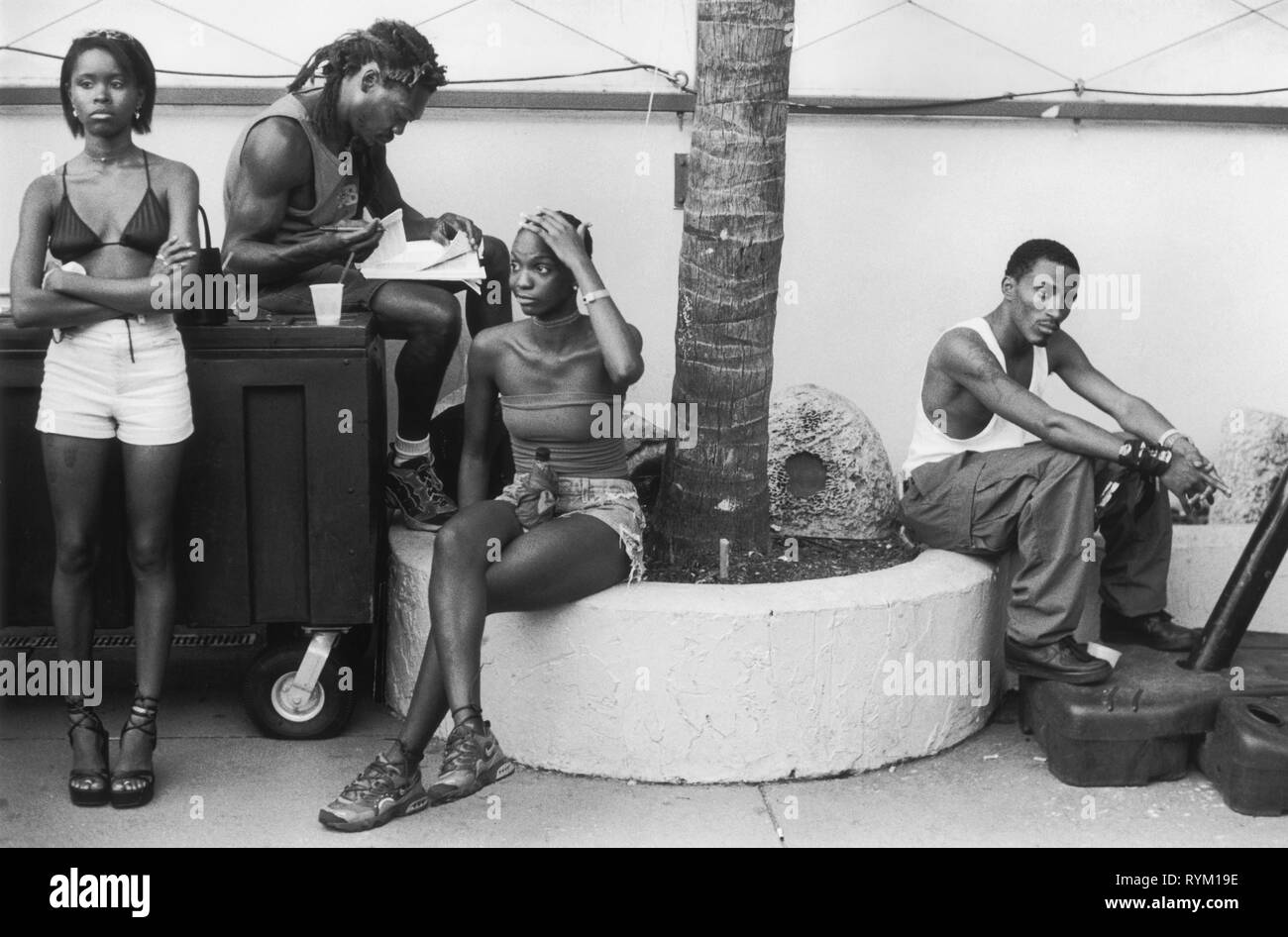 South Beach, Miami Florida USA 1990S.. Un gruppo di giovani uomini e donne afroamericani vestiti con pantaloncini corti e magliette si siedono e aspettano, assorbiti interamente dai propri pensieri. Un uomo fa qualche lavoro sui libri mentre un'altra donna tiene una bottiglia di vino in un sacchetto di carta tra le gambe. 1999 USA HOMER SYKES Foto Stock