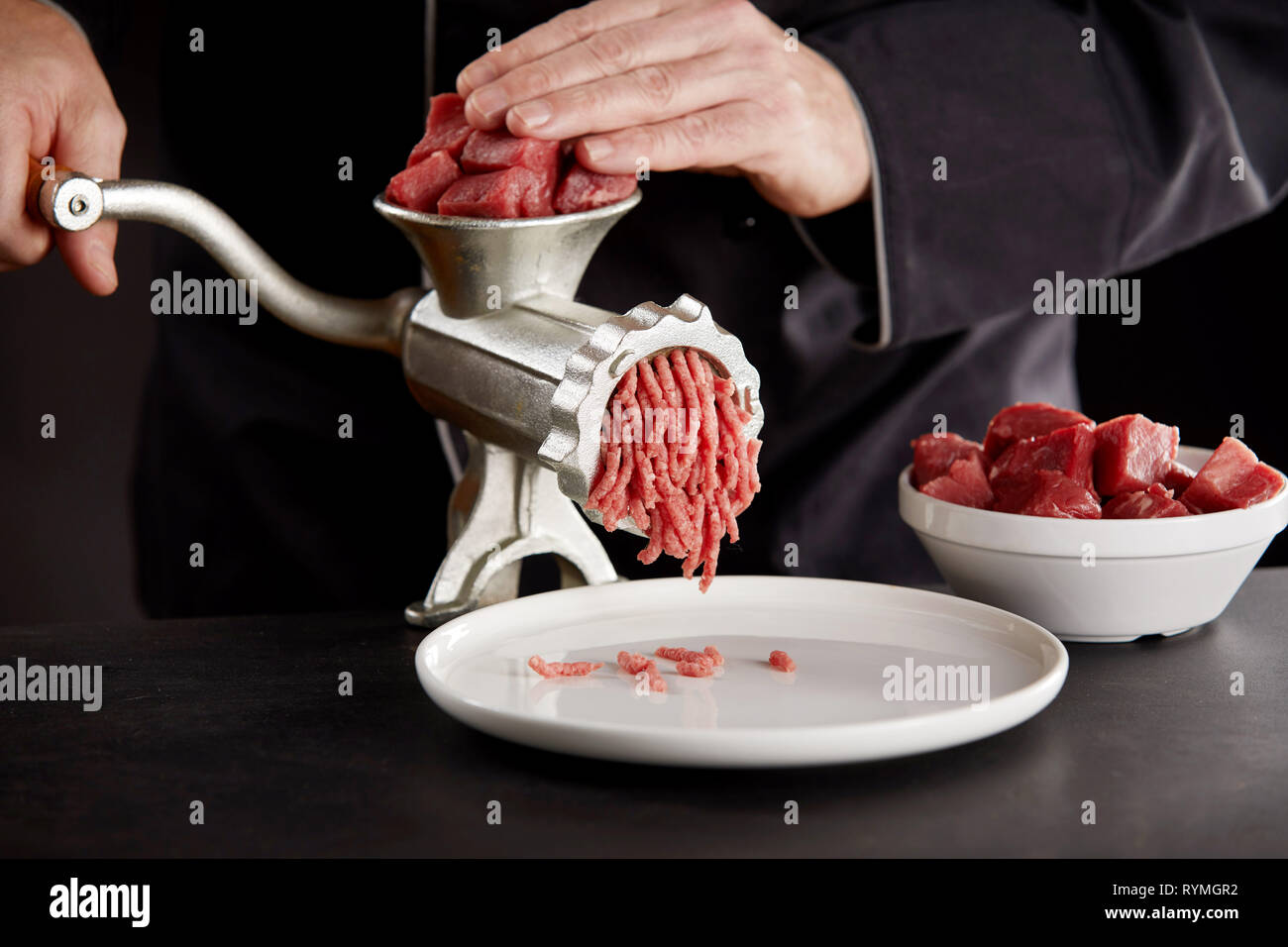 Cuocere in nero uniforme di macinazione delle carni bovine fresche o di maiale usando il vecchio manuale di metallo tritacarne sul banco di cucina. Pezzi di carni le carni rosse in bianco ciotola. Cooki Foto Stock