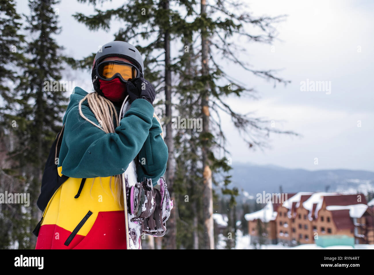 Ritratto di giovane donna con uno scarpone da snowboard su un pendio nevoso, base turistica in background. Foto Stock