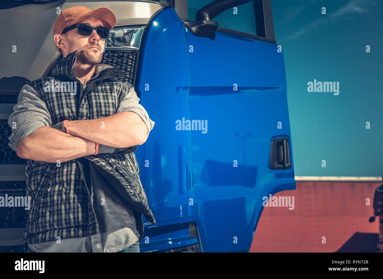 Trucker nella sua 30s indossando occhiali da sole e cappello da baseball nella parte anteriore del moderno semi carrello. Industria dei Trasporti lavoro. Foto Stock