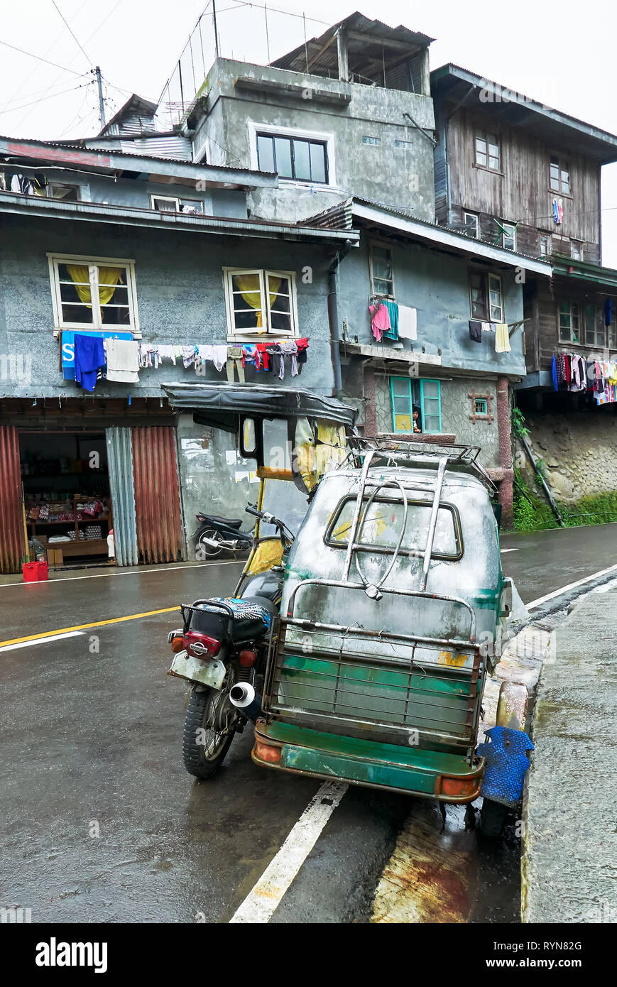 Un motociclo con sidecar parcheggio lungo una strada curva in Banaue Città, Provincia di Ifugao, Filippine, con i tipici edifici semplici in background. Foto Stock