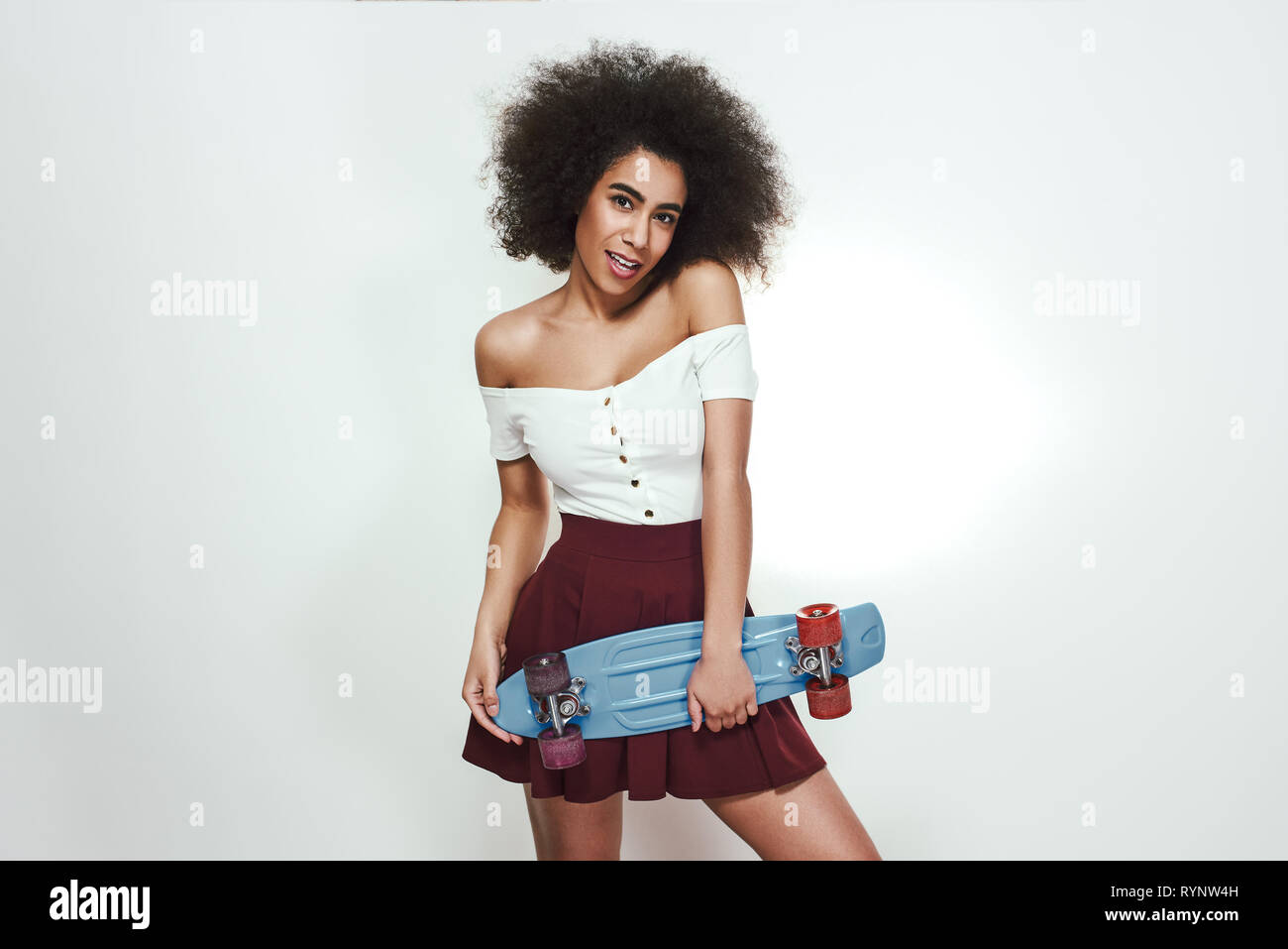 Io amo il pattinaggio! Studio shot della giovane e bella donna africana in abbigliamento casual holding skateboard e sorridere mentre in piedi contro uno sfondo grigio. Uno stile di vita attivo. Sport concept Foto Stock