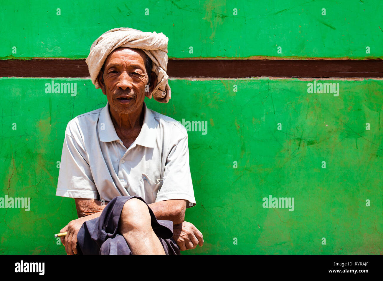Un tradizionale birmana l uomo si siede di fronte a un muro verde a Kalaw, Myanmar Foto Stock