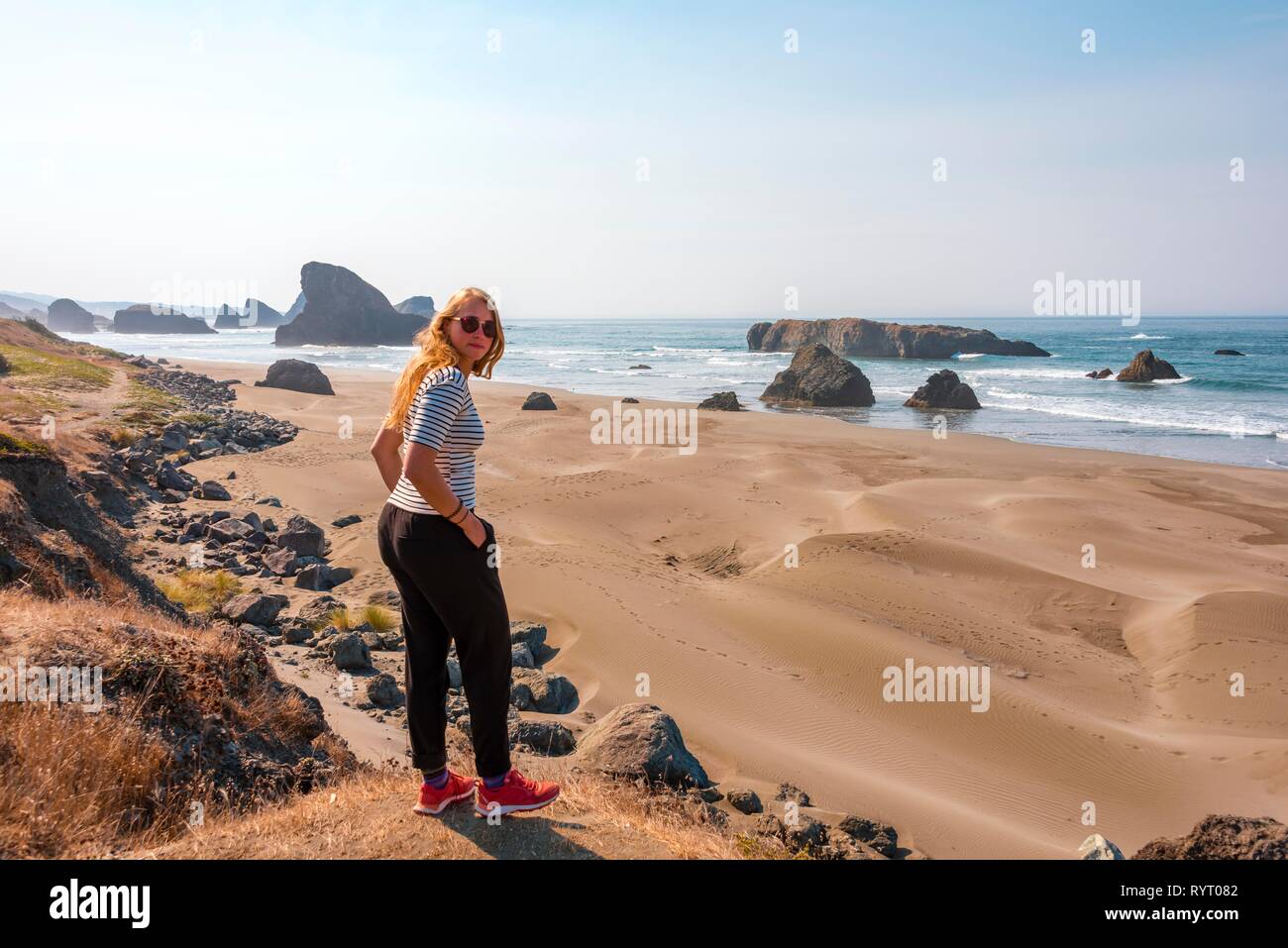 Giovane donna che guarda verso la fotocamera, spiaggia di sabbia e la costa con aspre rocce, Myers Creek Beach Viewpoint, Oregon, Stati Uniti d'America Foto Stock