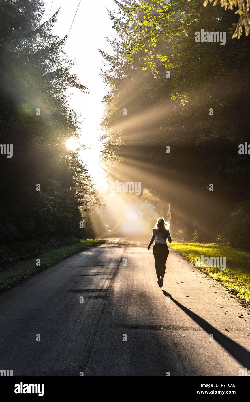 Giovane donna jogging lungo una strada, la luce del sole splende attraverso gli alberi, Oregon Coast Highway, Oregon, Stati Uniti d'America Foto Stock