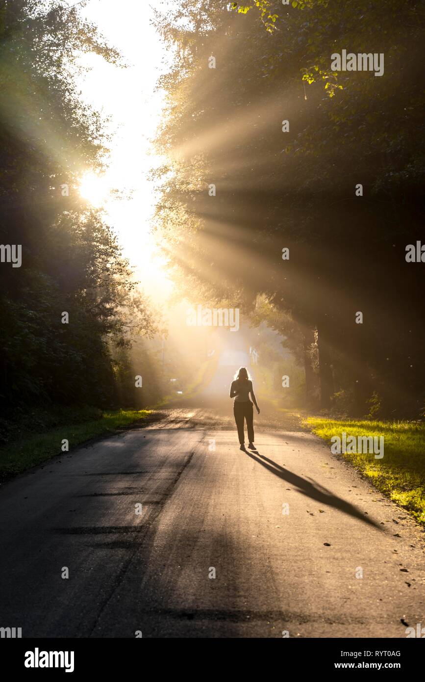 Giovane donna a camminare su una strada, la luce del sole che splende attraverso gli alberi, Oregon Coast Highway, Oregon, Stati Uniti d'America Foto Stock