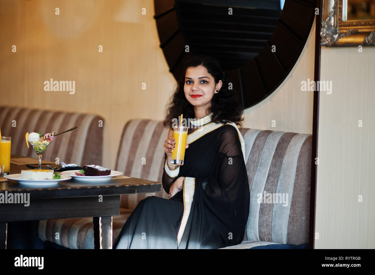 Giovane donna indiana usura elegante nera saree udienza del ristorante con bicchiere di succo di frutta a portata di mano. Foto Stock