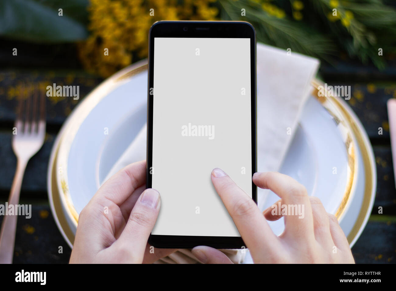 Mani tenendo lo smartphone su un tavolo con fiori e tabella ware Foto Stock