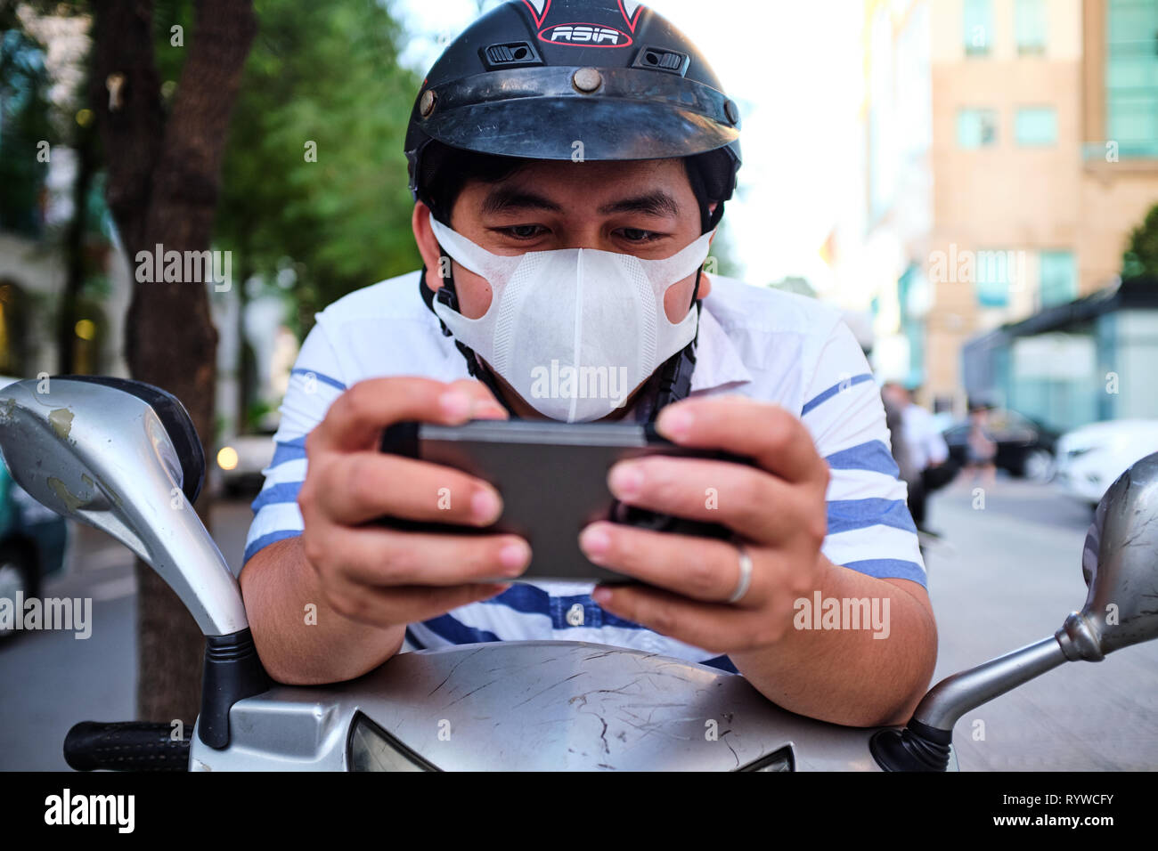 Nella foto è un uomo vietnamita di indossare una maschera sul suo scooter fermo controllo fuori il suo telefono Foto Stock