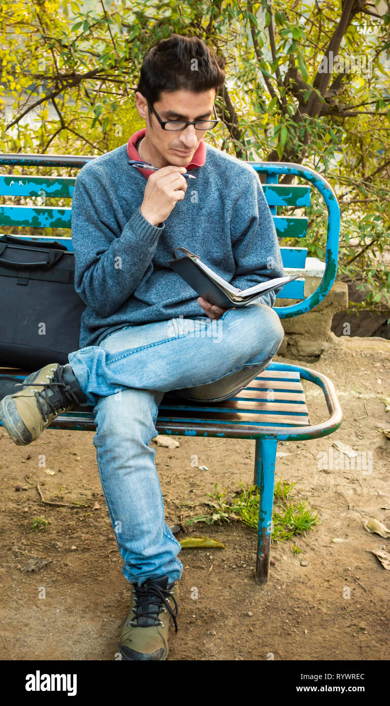 Immagine esterna di asiatici, indiani serena adulto giovane imprenditore lettura business planner e seduto su una panchina nel parco a tempo di giorno. Foto Stock