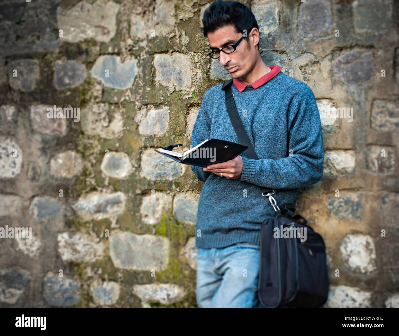 Immagine esterna di asiatici, indiani serena adulto giovane imprenditore lettura business planner e in piedi con il supporto della parete a tempo di giorno. Foto Stock