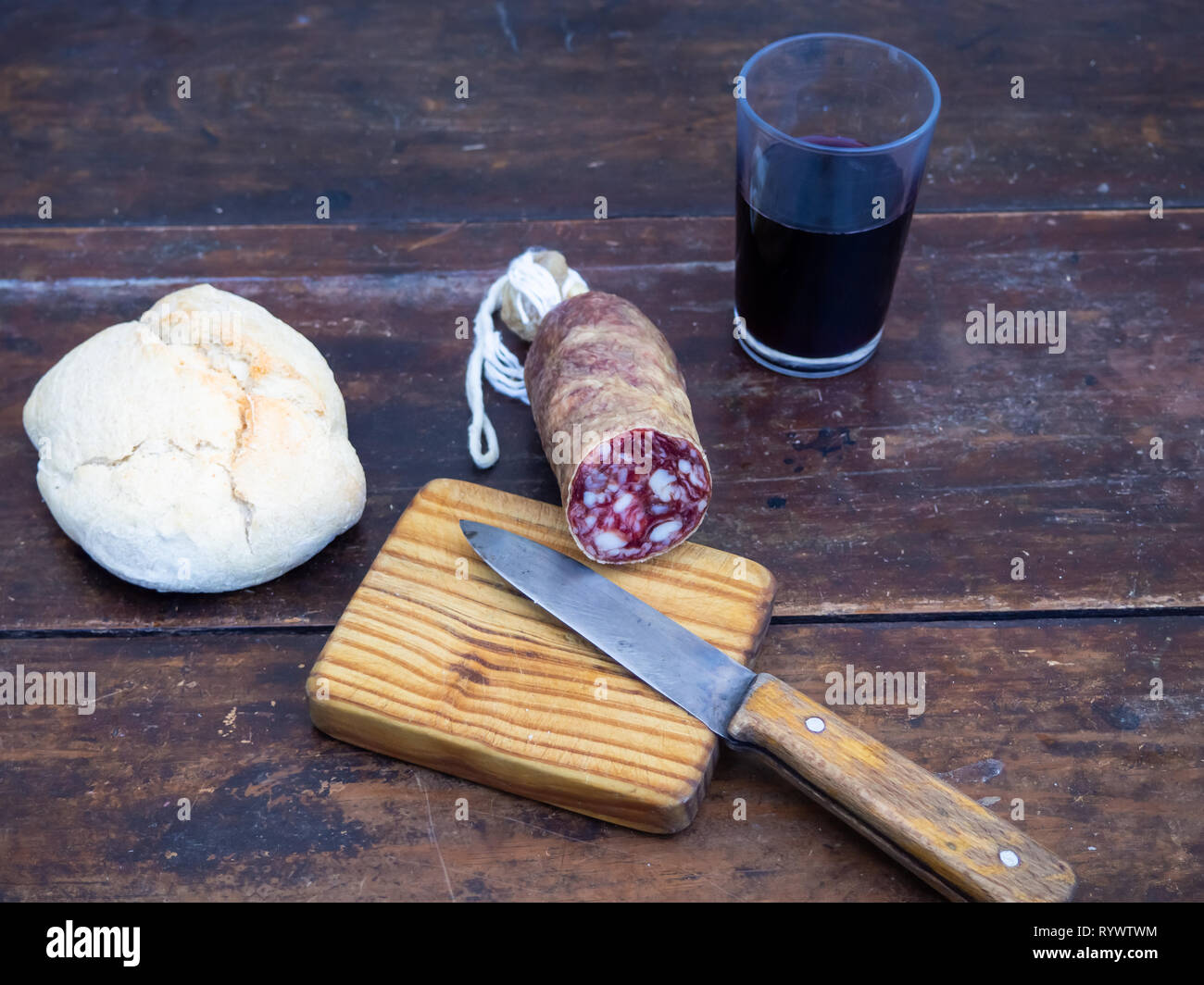 Un maiale iberico insaccato su una vecchia tavola di legno con un cuneo di formaggio di pecora, un coltello di antiquariato e una bottiglia e un bicchiere di rosso vino spagnolo Foto Stock