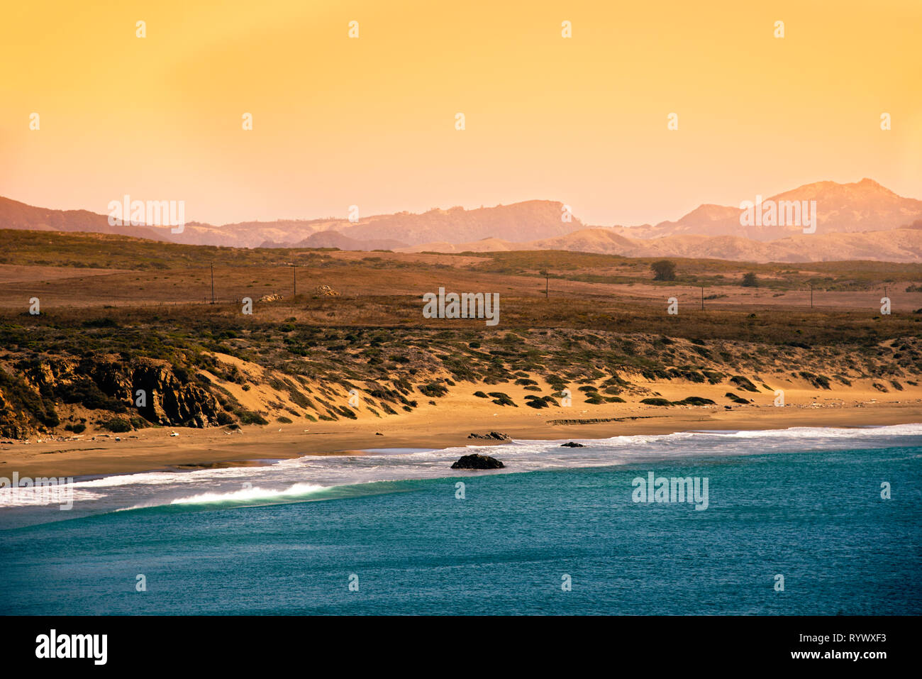 Opaco giallo arancione pomeriggio cielo sopra le montagne nebuloso e la spiaggia di sabbia fine e dorata con blu oceano. Onde che si infrangono sulla spiaggia. Foto Stock