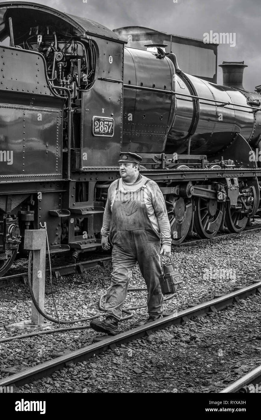 Vista laterale in bianco e nero della locomotiva a vapore del Regno Unito d'epoca stazionaria sulla ferrovia storica come autista del treno, in tuta e berretto passeggia per cambiare lampada. Foto Stock